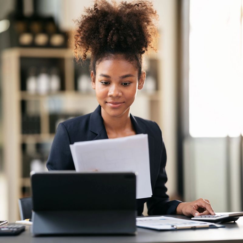 African american businesswoman in suit reading accounting information of business in paperwork and calculating accounting of new business on calculator while working in modern workspace.