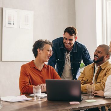 Four colleagues happily collaborate around a laptop in a bright office.
