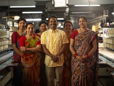 A group of six smiling factory workers standing together inside a textile mill.