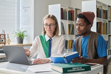 Two students studying English as a second language using a laptop and books.
