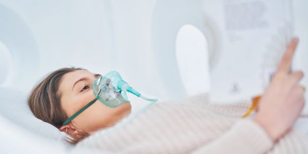 A woman wearing an oxygen mask lies in a hospital bed reading a document.