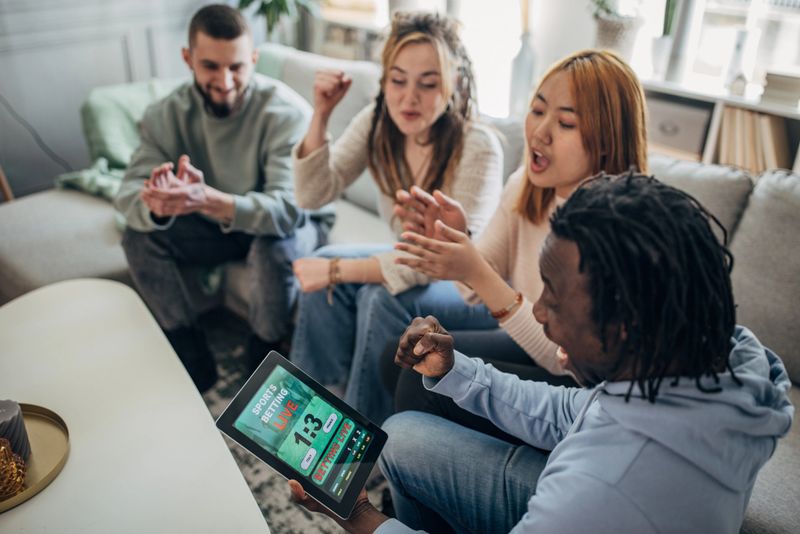 Multiracial group of people, friends gambling on a soccer match, using digital tablet while sitting in living room on sofa at home.