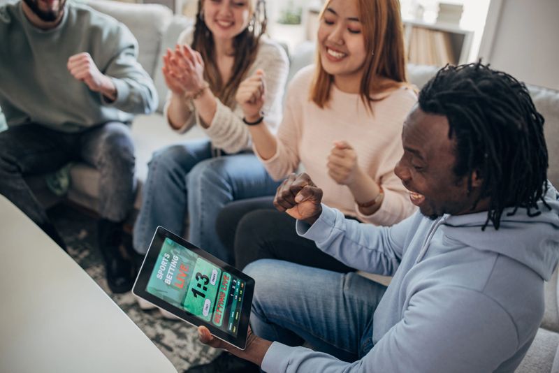 Multiracial group of people, friends gambling on a soccer match, using digital tablet while sitting in living room on sofa at home.