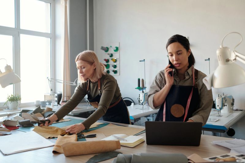 Young brunette female artisan using laptop and talking on smartphone by workplace while her mature colleague working with leather textile