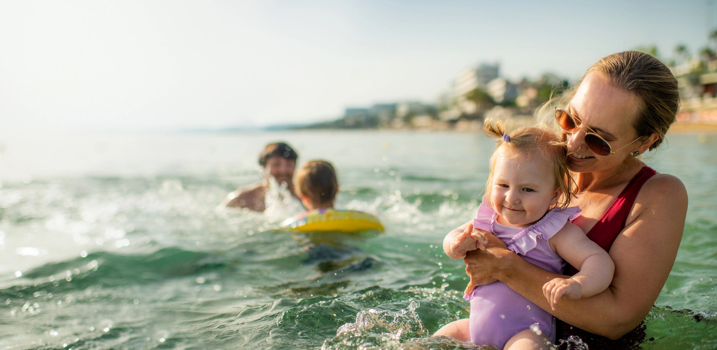Mother holding baby in the sea, family playing in the background.