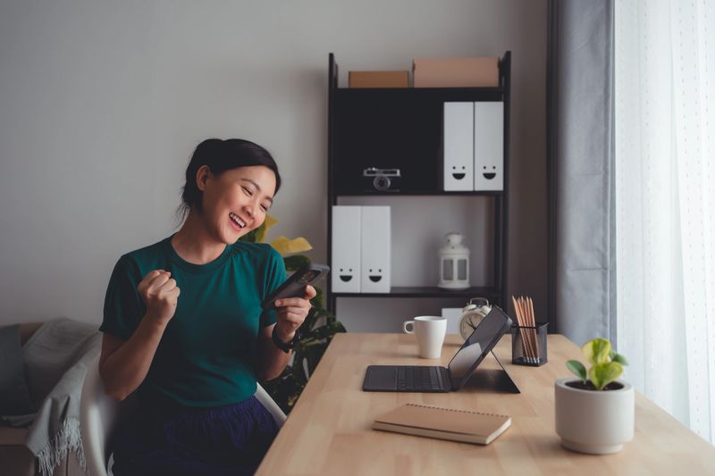 Asian woman holding and looking at smartphone happy excited and making a winning gesture sitting at home office.