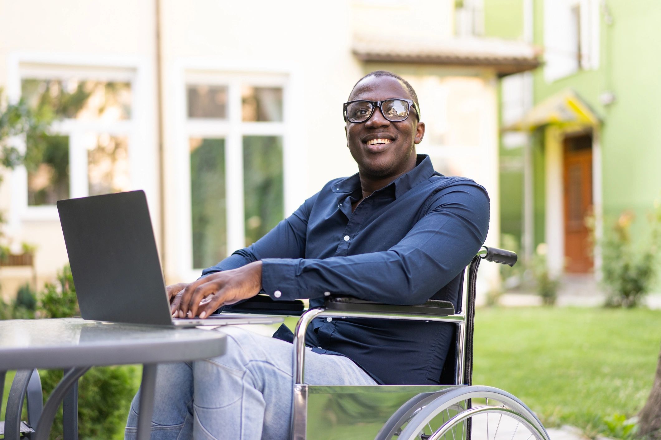 A man in a wheelchair works on a laptop at an outdoor table, smiling, with buildings and greenery in