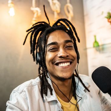 Smiling young man with dreadlocks and headphones speaking into a microphone.