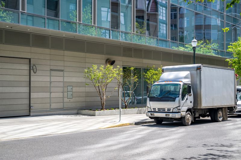 Industrial light duty transportation cab over powerful compact gray rig semi truck with big box trailer standing on the urban city street making the local delivery for different customers requests