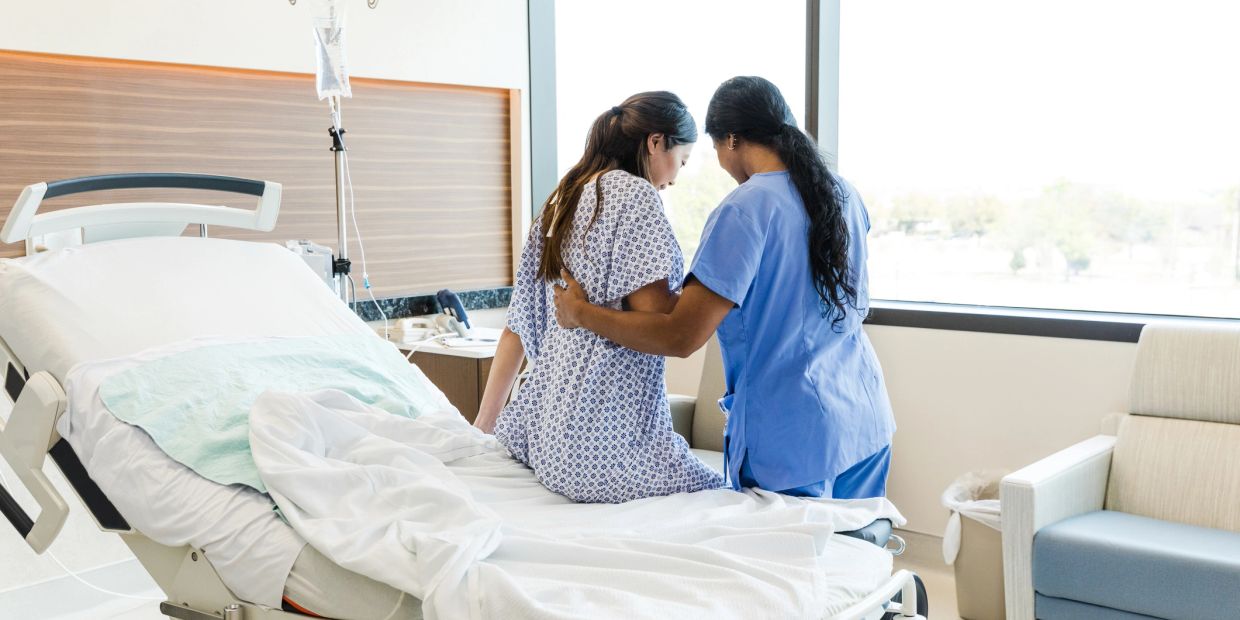 Nurse supporting a patient sitting on a hospital bed near a window.