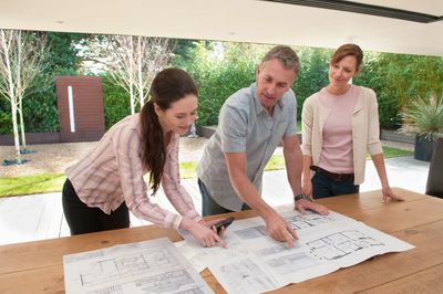 Three people discussing architectural plans at a table in a bright, outdoor setting.