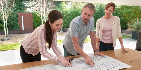 Three adults discussing architectural plans laid out on a table outdoors.