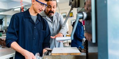 Instructor guiding student on machine operation in workshop.