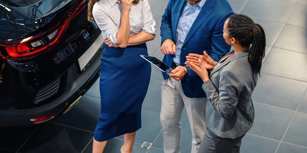 Three professionals discussing near a car, one holding a tablet.