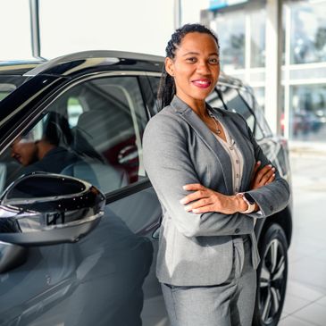 Confident woman in business suit standing by a car in a showroom.