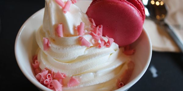 A cup of soft serve ice cream topped with pink chocolate shavings and a red macaron.
