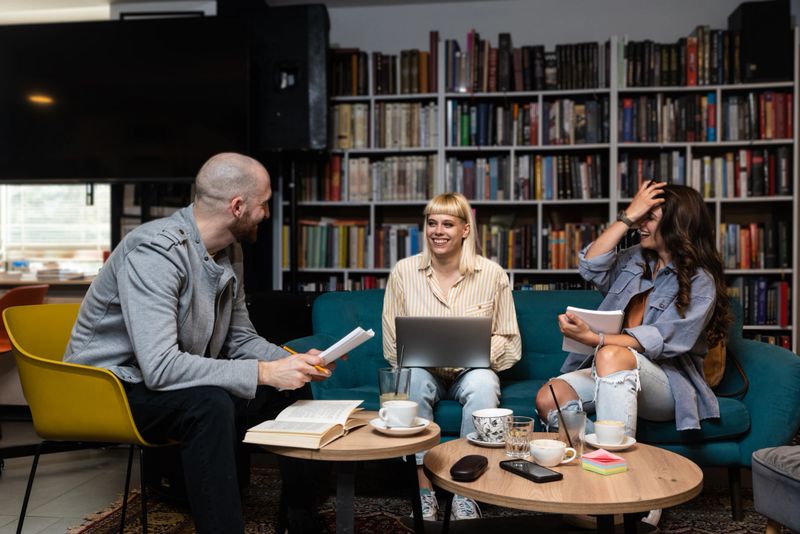 Group of three gen Z university students working together at laptop, writing notes, watching learning webinar, online lecture, reading tutorials on internet in college library. Education concept