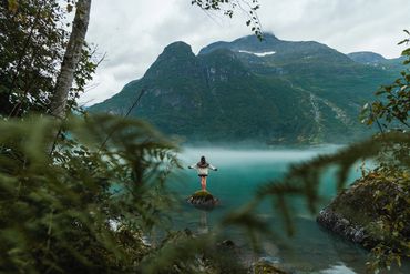 Person standing on a rock in a misty lake surrounded by mountains and trees.