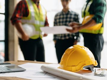 Yellow construction helmet on desk with engineers discussing plans in background.