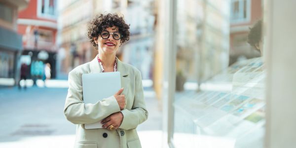 Smiling person with curly hair and glasses holding a laptop outdoors.