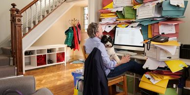 This image shows a person at a desk at home surrounded by piles of papers.