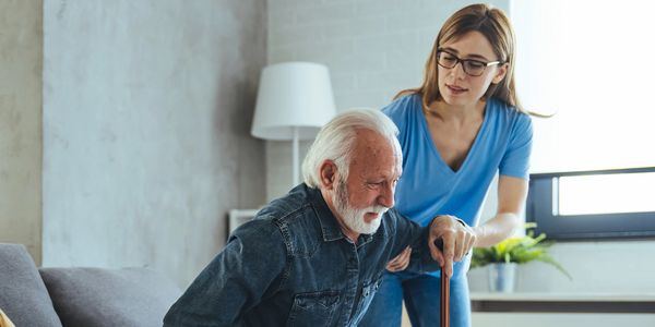 A caregiver assists an elderly man with a cane as he stands up from a couch.