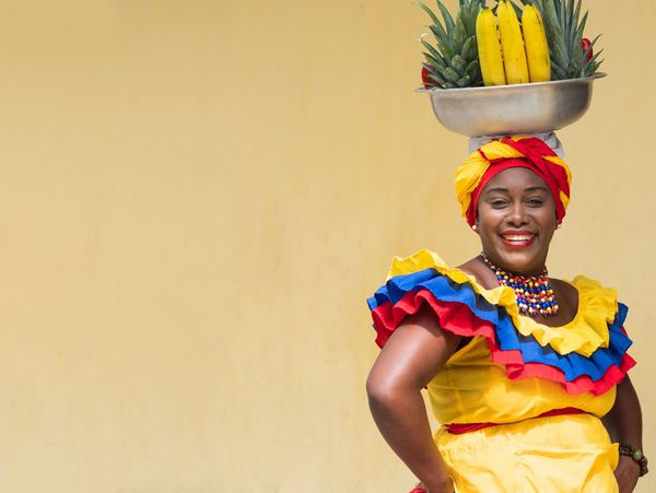 Smiling woman in colorful traditional attire with fruit basket on her head.