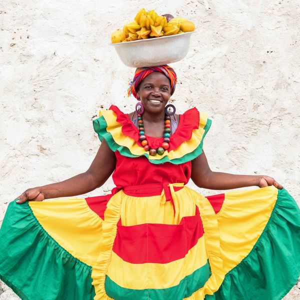 Smiling woman in colorful traditional dress balancing a bowl of fruit on her head.