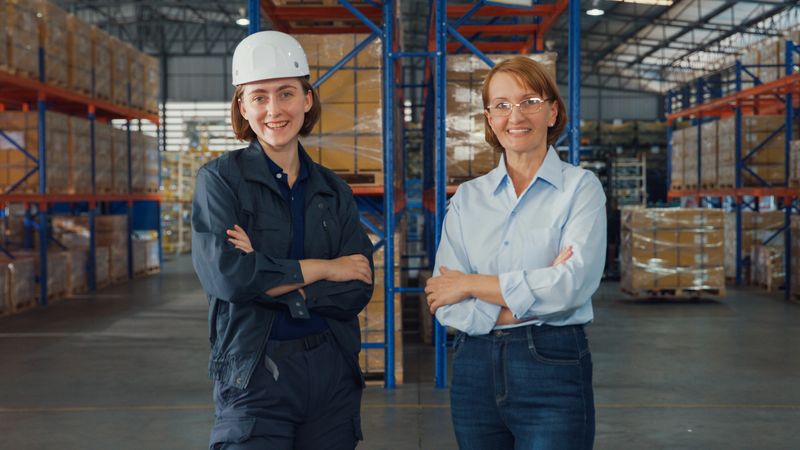 Young caucasian woman warehouse worker and mature female manager arms crossed, smiling and looking at camera in factory warehouse. Logistic industry business concept.
