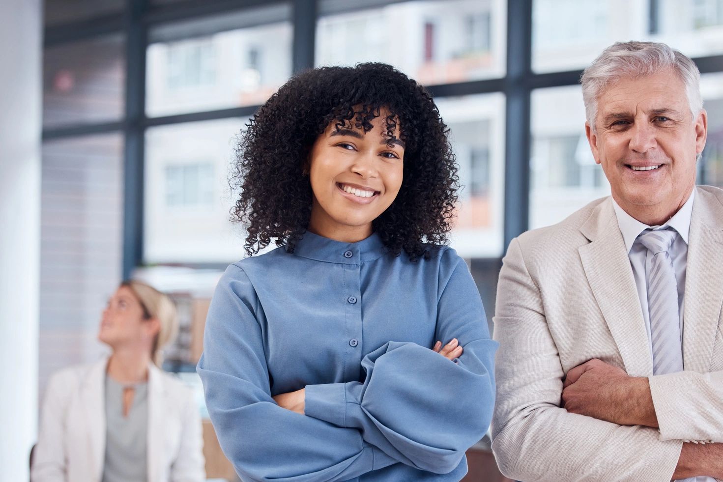 Two confident colleagues posing in a modern office with a team working behind them.
