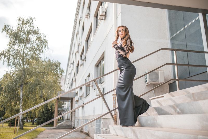Beautiful modern woman in black long dress with bare back standing outdoors and posing for camera