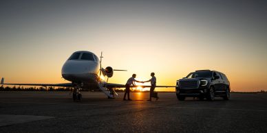 Two people shaking hands near a private jet and SUV at sunset.