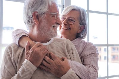 Elderly woman embraces her elderly husband from behind while they both smile. 