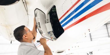 Technician inspecting an open panel on an aircraft fuselage in a hangar.