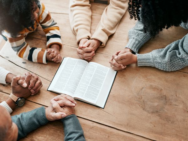 Group of people holding hands around an open book in prayer.