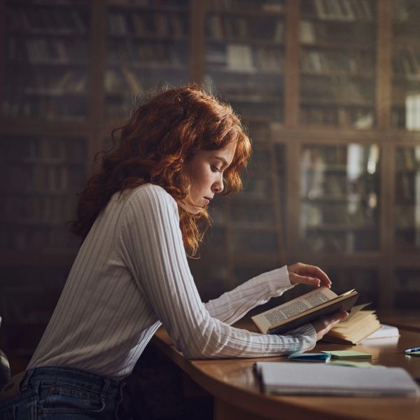 A young woman with red hair reading a book at a library desk.