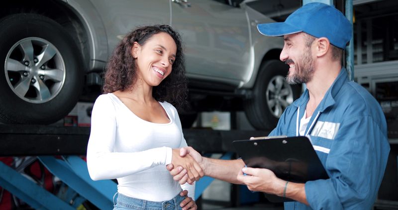 Happy automotive mechanic man in uniform shaking hands with women client at auto repair shop. Car service, repair, maintenance, gestures and man-mechanic concept