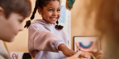 A smiling girl reaching out to give a fist bump in a classroom setting.