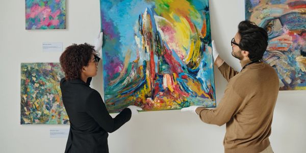 Two people hanging abstract paintings in a gallery.