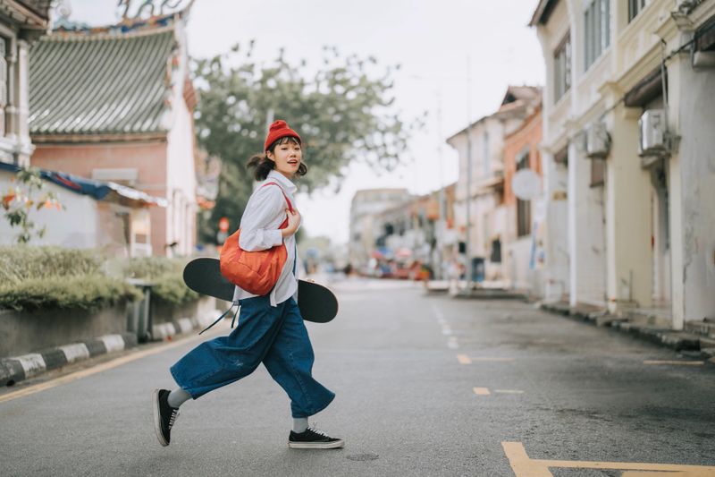 Happy Asian Chinese young woman crossing road carrying skateboard in old town