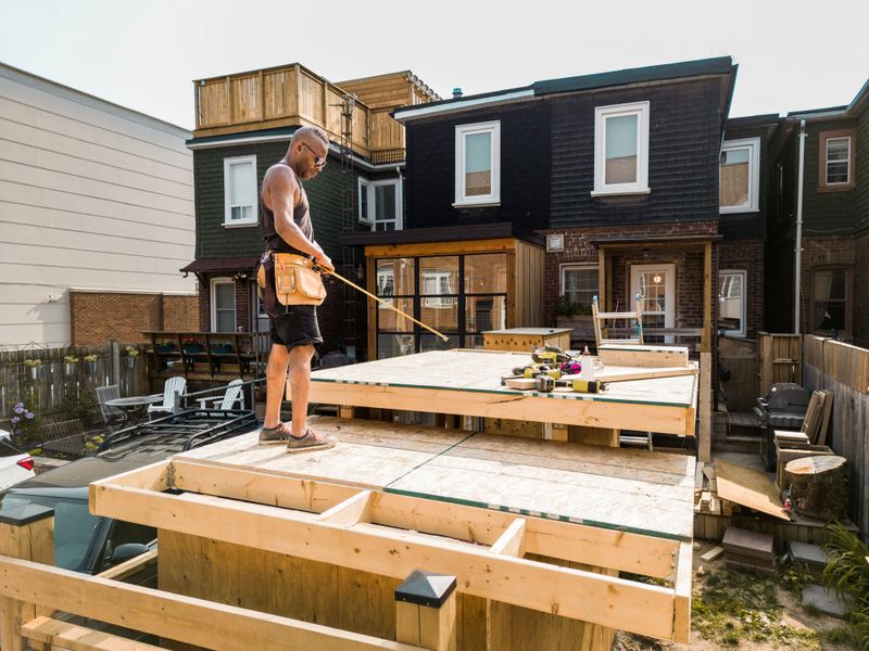 Black Male carpenter building shed in the backyard. He is dressed in summer work clothes. Exterior of urban back yard during the day. Captured by drone.