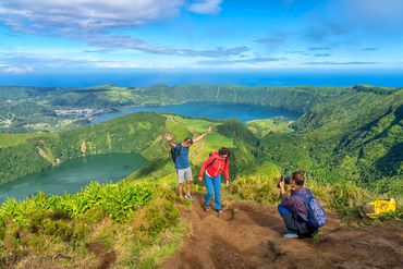 Hikers capturing scenic views atop a lush mountain with lakes and ocean in the background.