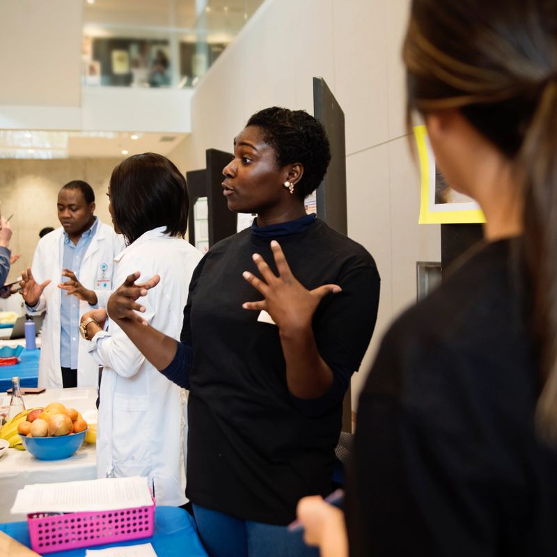 Young adult female college student in science fair. They are standing at their kiosk, explaining their work. Another group of student in the background. They are from the medical department. Square waist up indoors shot with copy space. This was taken in Quebec, Canada.
