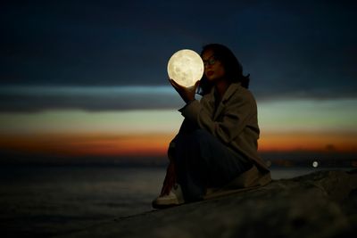 Woman holding a glowing moon prop at twilight by the water.