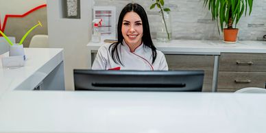Smiling receptionist at a modern white counter with plants and a computer.