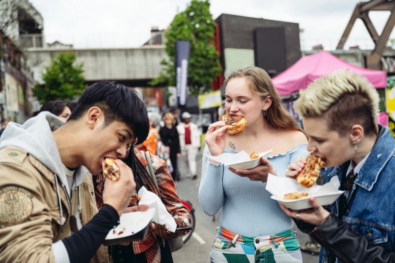 Waist-up view of four Londoners in casual clothing standing together outdoors and taking bites of delicious grilled sandwiches.