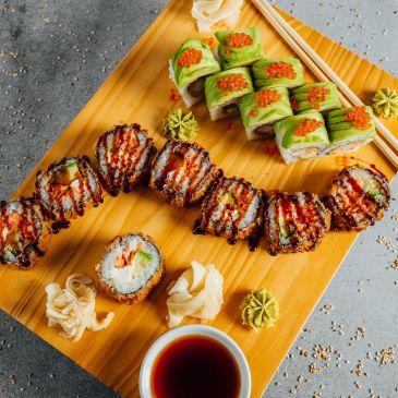 Assorted sushi rolls with avocado, sauce, and garnishes on a wooden board.