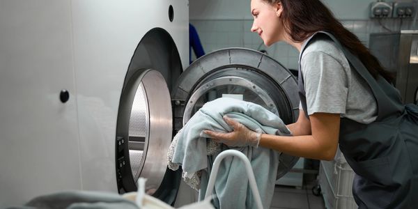 A woman loads laundry into a washing machine, wearing gloves and a gray apron.