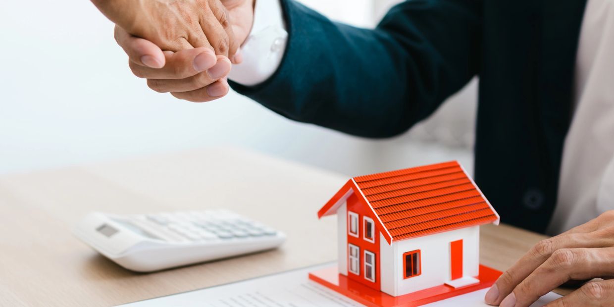 Handshake over a small house model and calculator on desk.