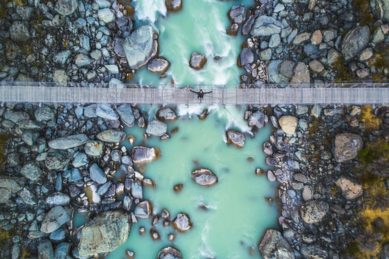 Looking down on young happy man on suspension bridge over glacial river in the mountains. Photographed in New Zealand.